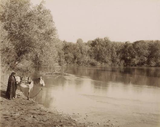 RECORD DATE NOT STATED Cours du Jourdain.- The course of the Jordan, Man with a horse on the bank of the Jordan, photograph, Part of Travel album with photos of Jerusalem and other biblical places., negative number: 916 , photographer: Maison Bonfils, Jordan (river), c. 1867 - c. 1895, pasteboard, paper, albumen print, height 227 mm x width 284 mm, riding a horse, ass, or mule, rider, horseman, river (landscape with figures, staffage), A riverside scene with calm reflective water edged by a tree-lined far bank and a large leafy tree on the near left, a person standing at the water s edge holding a rope attached to a saddled horse that faces the water, the person wearing a long dark garment and a head covering and leaning on a long staff, vegetation and low shrub Copyright Penta Springs LCK260531_18262