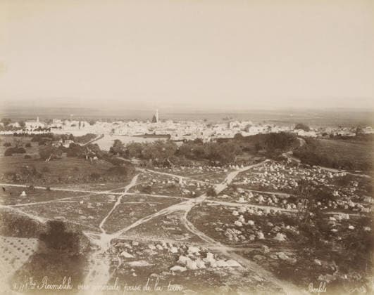 RECORD DATE NOT STATED View of Ramla in Israel, Ramallah and surrounding land, seen from a tower (former title), Rameleh, vue gà nà rale prise de la tour, photograph, View of Ramla with the Plain of Sharon and the Mountains of Judea, in the foreground an Islamic cemetery. Part of Travel Album with photographs of Jerusalem and other biblical places., negative number: 241bis , photographer: Maison Bonfils, Ramallah, c. 1867 - c. 1895, Palestine, pasteboard, paper, albumen print, height 225 mm x width 283 mm, prospect of city, town panorama, silhouette of city, Ramla, A sepia-toned aerial view shows a broad landscape with a foreground of rocky, uneven ground marked by winding dirt paths and scattered stone markers forming small rectangular shapes, some grouped and Copyright Penta Springs LCK260531_18265