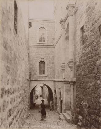 RECORD DATE NOT STATED VI Station, maison de Ste VÃ ronique, Sixth Stations of the Cross on the Via Dolorosa in Jerusalem, album page, photograph, This photo is part of an album., number: 265 , photographer: FÃ lix Bonfils, Jerusalem, 1867 - 1885, paper, albumen print, height 277 mm x width 219 mm, street, Via Dolorosa., A narrow cobblestone alley flanked by tall stone buildings with textured walls and arched openings, a stone archway spanning the alley and supporting an upper-level building with shuttered and arched windows, ornate carved columns and decorative stone work on the right building, a few people standing and walking in the alley including a woman and child in the foreground and others further back beneath the arch, small steps and recessed doorways Copyright Penta Springs LCK260618_00807