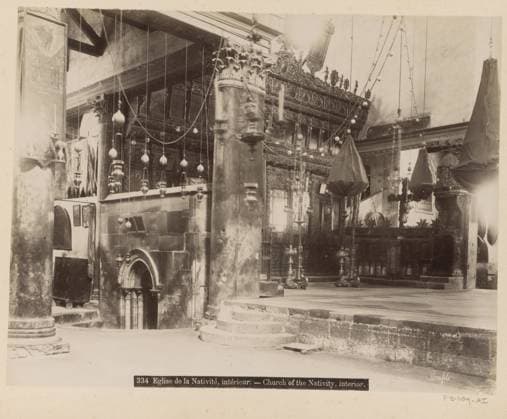 RECORD DATE NOT STATED Eglise de la Natività , intà rieur.- Church of the Nativity, interior., Interior of the Church of the Nativity in Bethlehem, photograph, Part of a travel album with photos of Jerusalem and other biblical places., negative number: 334 , photographer: Maison Bonfils, Bethlehem (Israel), c. 1867 - c. 1895, Palestine, pasteboard, paper, albumen print, height 217 mm x width 283 mm, interior of church., The image shows an interior space with stone columns and carved architectural details, including an arched doorway and stepped platforms. Hanging lamps and chandeliers descend from the ceiling, some with decorative bulbous shapes and others covered in draped fabric bags. Ornate carved wooden screens and panels with intricate patterns line the bac Copyright Penta Springs LCK260621_006369