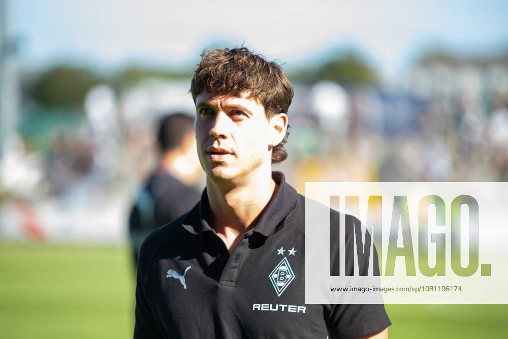 A Borussia Mönchengladbach official stands in front of the field wearing a black club polo shirt with the Puma logo and REUTER sponsorship. Spectators and the field can be seen in the background. Borussia Mönchengladbach – Official in Velbert
