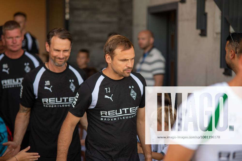 Two members of the Borussia Mönchengladbach coaching staff, wearing black practice shirts with REUTER sponsorship logos, walk through an entrance surrounded by fans and children. The initials TJ and JM are visible on the shirts. The Borussia Mönchengladbach coaching staff as they enter the field in Velbert