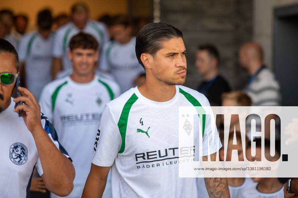 Kevin Diks (No. 4) of Borussia Mönchengladbach walks onto the field in Velbert with the team, looking focused. In the background, other Borussia Mönchengladbach players, spectators, and children can be seen. Kevin Diks (No. 4) of Borussia Mönchengladbach during the walkout in Velbert