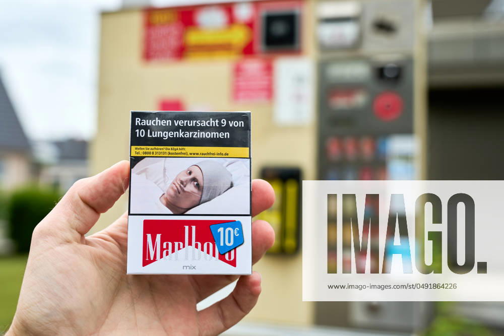 Hand holds a pack of Marlboro cigarettes costing 10 euros in front of a cigarette machine. There is a warning on the cigarettes: Smoking causes 9 out of 10 lung cancers