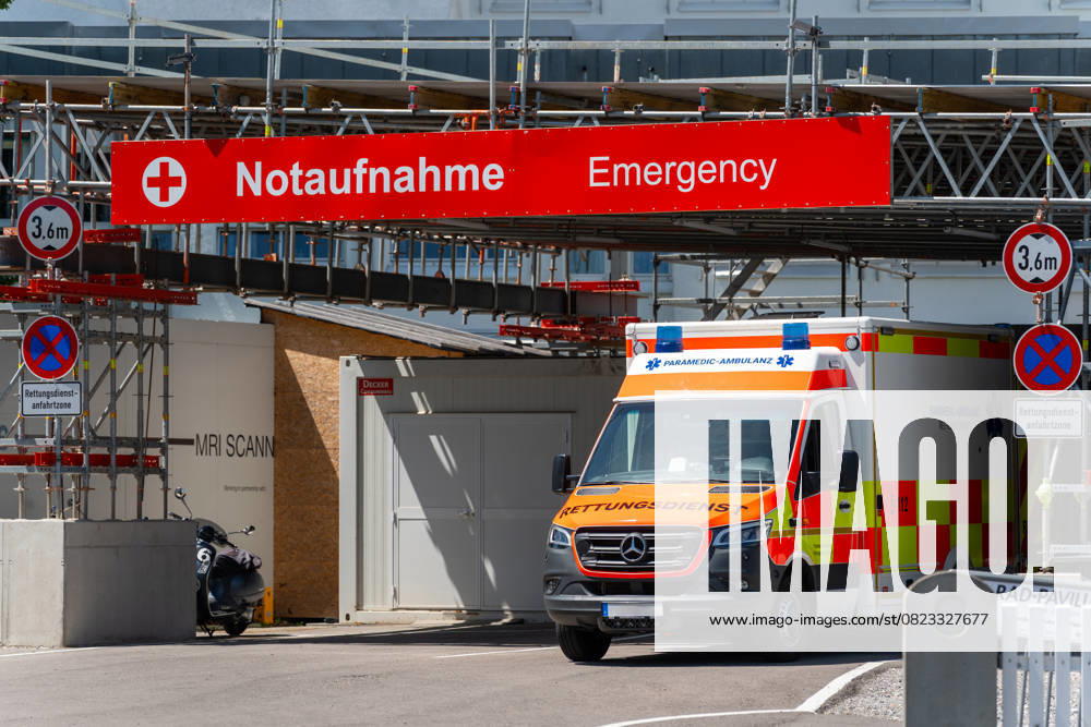 An ambulance stands in front of the entrance to an emergency room with clearly visible signage in German and English, in front of a modern hospital