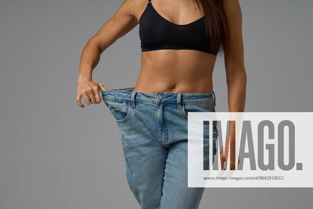 Fit Frau in schwarzem Sport-BH zieht lose Jeans, um Gewichtsverlust Fortschritte auf grauem Hintergrund zu zeigen Fit young woman wearing black sports bra and oversized jeans, pulling the waistband to display weight loss results. Studio shot on neutral gray backdrop. (License=RF) ,model released, Symbolfoto