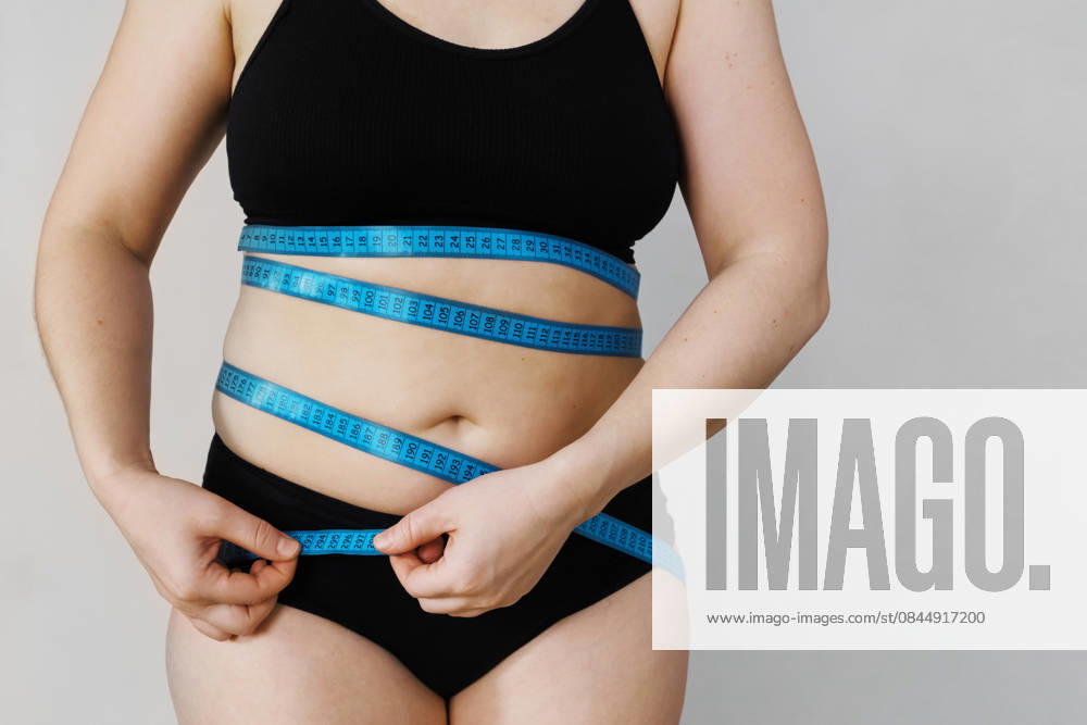 Eine Frau steht vor einem schlichten Hintergrund und misst ihre Taille mit einem blauen Maßband Eine Frau steht vor einem schlichten Hintergrund und misst ihre Taille mit einem blauen Maßband (License=RF) ,model released, Symbolfoto