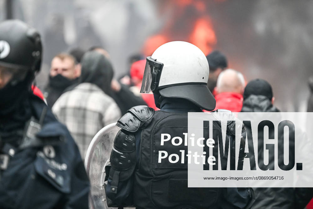 Heavily equipped police officers wearing helmets and carrying riot shields stand amid smoke and fire, with a blurred crowd in the background, near Mont des Arts in Brussels, Belgium, on 9 October 2026. The scene shows police deployed during violent confrontations and public disorder in the city centre.