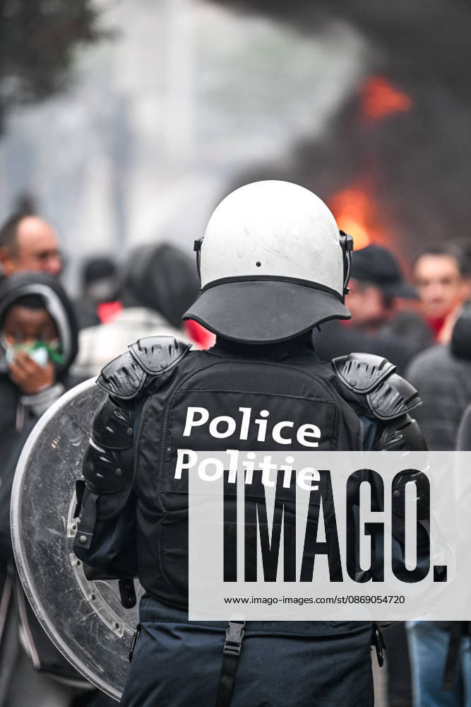 Heavily equipped police officers wearing helmets and carrying riot shields stand amid smoke and fire, with a blurred crowd in the background, near Mont des Arts in Brussels, Belgium, on 9 October 2026. The scene shows police deployed during violent confrontations and public disorder in the city centre.