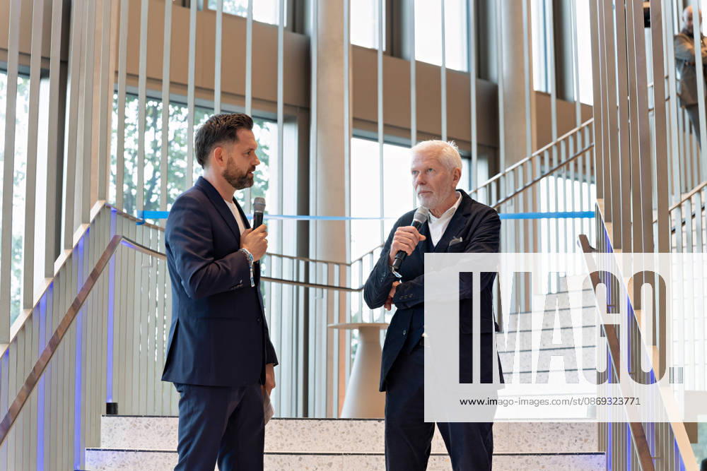 Berlin, 13.10.2026: Ein Moderator (l.) im Gespräch mit dem Architekten des Gebäudes vom Büro Kleihues + Kleihues (r.) auf der Treppe im Foyer des neuen DKB-Campus in der Europacity in Berlin-Mitte während der Eröffnungsfeier; im Hintergrund ist das blaue Band für den symbolischen Banddurchschnitt gespannt. Die Deutsche Kreditbank AG (DKB) hat ihren neuen Hauptsitz heute offiziell eröffnet. Der von Kleihues + Kleihues entworfene, rund 82 Meter hohe Komplex besteht aus drei Gebäudeteilen mit fünf, elf und 19 Geschossen und rund 35.000 Quadratmetern Mietfläche; entwickelt wurde er von der CA Immo unter dem Projektnamen Upbeat. Rund 2.600 Mitarbeitende des DKB-Konzerns, die bislang auf mehrere Berliner Standorte verteilt waren,