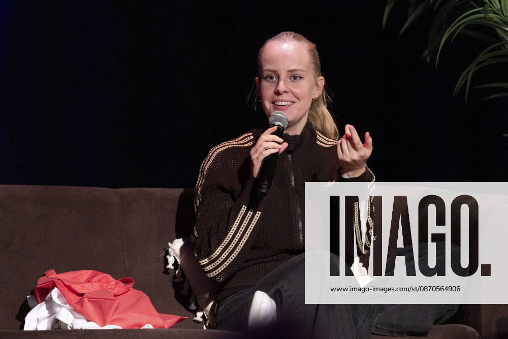 Poetry artist and author Clara LOESEL LÖSEL speaking, gesturing with one hand, during her reading—a preview event for her new book, *Ich dachte, sowas fühle nur ich*—sitting on a sofa with a microphone; single frame, single photograph, at the Theater am Tanzbrunnen, Cologne, on October 23, 2026