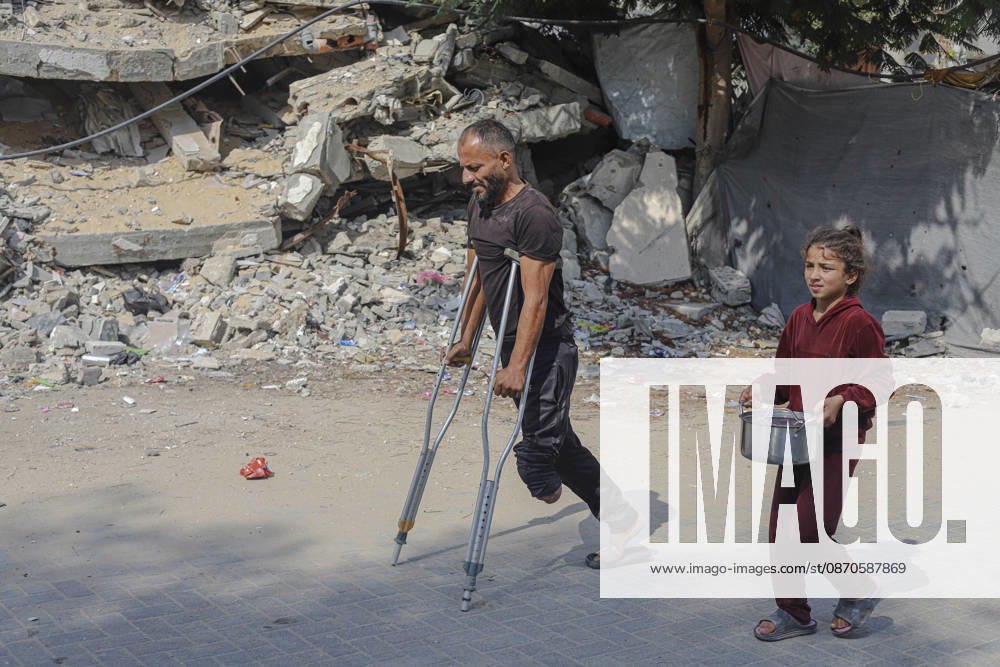 Gaza Palestinians Food Shortages 2026 A Palestinian man using crutches walks beside a young girl carrying a pot through a neighborhood of damaged buildings in Jabalia Refugee Camp, northern Gaza Strip, Palestine, on October 24, 2026. The UN says widespread damage, displacement and food shortages have left families across Gaza heavily dependent on humanitarian assistance, with food distributions reduced in October amid funding constraints. Jabalia Gaza Strip Palestinian Territories