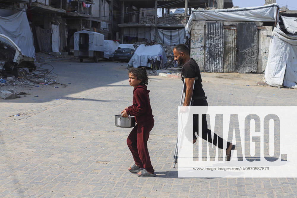 Gaza Palestinians Food Shortages 2026 A Palestinian man using crutches walks beside a young girl carrying a pot through a neighborhood of damaged buildings in Jabalia Refugee Camp, northern Gaza Strip, Palestine, on October 24, 2026. The UN says widespread damage, displacement and food shortages have left families across Gaza heavily dependent on humanitarian assistance, with food distributions reduced in October amid funding constraints. Jabalia Gaza Strip Palestinian Territories