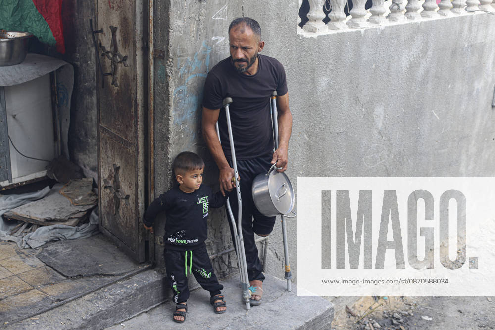 Gaza Palestinians Food Shortages 2026 A Palestinian man using crutches stands with a young child outside a damaged building while holding a metal cooking pot in Jabalia Refugee Camp, northern Gaza Strip, Palestine, on October 24, 2026. The UN says widespread damage, displacement and food shortages have left families across Gaza heavily dependent on humanitarian assistance, with food distributions reduced in October amid funding constraints. Jabalia Gaza Strip Palestinian Territories