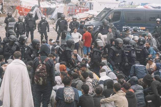 October 4, 2026, Ceuta, Ceuta, Spain: Riot police officers stand among migrants sitting on the ground, one wrapped in a blanket, during the eviction of the makeshift camp on El Trampolin beach in Ceuta. Spanish National Police began evicting the makeshift camp on El Trampolin beach in Ceuta on October 4, where more than 3,000 migrants, most of them young Moroccan men, were living. Dozens of police vans, armoured vehicles of the Police Intervention Units (UIP), drones and boats took part in the operation. People were taken by bus to the temporary centre at the Tarajal border to be identified, and the authorities plan to clear the beach in phases before October 15. The camp grew after a mass arrival at the end of July 2026 set off a migration - ZUMAs392 20261004_zip_s392_023 Copyright: xFabianxSuspensivox