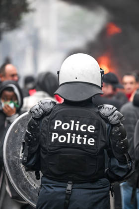 Heavily equipped police officers wearing helmets and carrying riot shields stand amid smoke and fire, with a blurred crowd in the background, near Mont des Arts in Brussels, Belgium, on 9 October 2026. The scene shows police deployed during violent confrontations and public disorder in the city centre.
