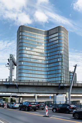 Berlin, 13.10.2026: Autos stehen im Stau auf einer Straße unterhalb einer Bahnbrücke, dahinter ragt das Hochhaus des neuen DKB-Campus in der Europacity in Berlin-Mitte auf. Die Deutsche Kreditbank AG (DKB) hat ihren neuen Hauptsitz heute offiziell eröffnet. Der von Kleihues + Kleihues entworfene, rund 82 Meter hohe Komplex besteht aus drei Gebäudeteilen mit fünf, elf und 19 Geschossen und rund 35.000 Quadratmetern Mietfläche; entwickelt wurde er von der CA Immo unter dem Projektnamen Upbeat. Rund 2.600 Mitarbeitende des DKB-Konzerns, die bislang auf mehrere Berliner Standorte verteilt waren, arbeiten nun gemeinsam im Campus. Die DKB ist Exklusivmieterin und hat einen Mietvertrag über 15 Jahre abgeschlossen. DKB eröffnet neuen