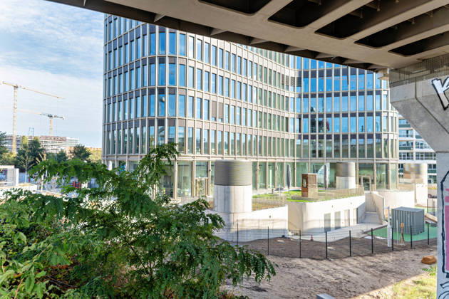 Berlin, 13.10.2026: Blick unter einer Brücke hindurch auf den Sockel des neuen DKB-Campus in der Europacity in Berlin-Mitte mit begrünten Terrassen; links drehen sich Baukräne im weiter wachsenden Stadtquartier. Die Deutsche Kreditbank AG (DKB) hat ihren neuen Hauptsitz heute offiziell eröffnet. Der von Kleihues + Kleihues entworfene, rund 82 Meter hohe Komplex besteht aus drei Gebäudeteilen mit fünf, elf und 19 Geschossen und rund 35.000 Quadratmetern Mietfläche; entwickelt wurde er von der CA Immo unter dem Projektnamen Upbeat. Rund 2.600 Mitarbeitende des DKB-Konzerns, die bislang auf mehrere Berliner Standorte verteilt waren, arbeiten nun gemeinsam im Campus. Die DKB ist Exklusivmieterin und hat einen Mietvertrag über 15 J