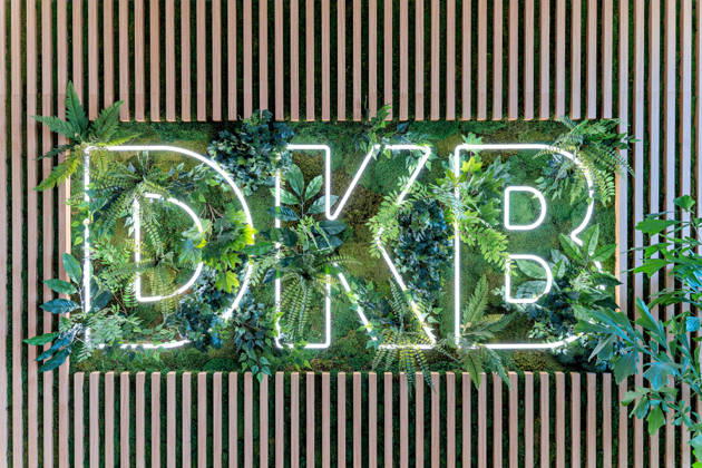 Berlin, 13.10.2026: Das DKB-Logo als Leuchtschrift in einer Pflanzenwand mit Moos und Holzlamellen im neuen DKB-Campus in der Europacity in Berlin-Mitte. Die Deutsche Kreditbank AG (DKB) hat ihren neuen Hauptsitz heute offiziell eröffnet. Der von Kleihues + Kleihues entworfene, rund 82 Meter hohe Komplex besteht aus drei Gebäudeteilen mit fünf, elf und 19 Geschossen und rund 35.000 Quadratmetern Mietfläche; entwickelt wurde er von der CA Immo unter dem Projektnamen Upbeat. Rund 2.600 Mitarbeitende des DKB-Konzerns, die bislang auf mehrere Berliner Standorte verteilt waren, arbeiten nun gemeinsam im Campus. Die DKB ist Exklusivmieterin und hat einen Mietvertrag über 15 Jahre abgeschlossen. Zum Campus gehören neben modernen Arbeit