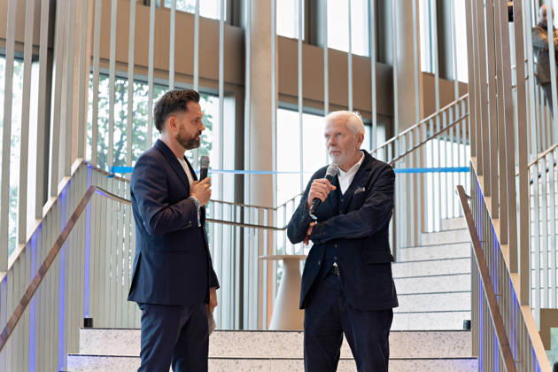 Berlin, 13.10.2026: Ein Moderator (l.) im Gespräch mit dem Architekten des Gebäudes vom Büro Kleihues + Kleihues (r.) auf der Treppe im Foyer des neuen DKB-Campus in der Europacity in Berlin-Mitte während der Eröffnungsfeier; im Hintergrund ist das blaue Band für den symbolischen Banddurchschnitt gespannt. Die Deutsche Kreditbank AG (DKB) hat ihren neuen Hauptsitz heute offiziell eröffnet. Der von Kleihues + Kleihues entworfene, rund 82 Meter hohe Komplex besteht aus drei Gebäudeteilen mit fünf, elf und 19 Geschossen und rund 35.000 Quadratmetern Mietfläche; entwickelt wurde er von der CA Immo unter dem Projektnamen Upbeat. Rund 2.600 Mitarbeitende des DKB-Konzerns, die bislang auf mehrere Berliner Standorte verteilt waren,
