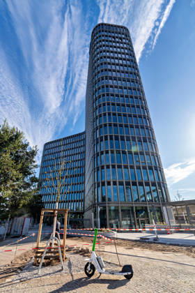 Berlin, 13.10.2026: Der rund 82 Meter hohe Turm des neuen DKB-Campus in der Europacity in Berlin-Mitte; im Vordergrund ein frisch gepflanzter Baum und ein abgestellter E-Scooter, die Außenanlagen sind noch nicht ganz fertiggestellt. Die Deutsche Kreditbank AG (DKB) hat ihren neuen Hauptsitz heute offiziell eröffnet. Der von Kleihues + Kleihues entworfene, rund 82 Meter hohe Komplex besteht aus drei Gebäudeteilen mit fünf, elf und 19 Geschossen und rund 35.000 Quadratmetern Mietfläche; entwickelt wurde er von der CA Immo unter dem Projektnamen Upbeat. Rund 2.600 Mitarbeitende des DKB-Konzerns, die bislang auf mehrere Berliner Standorte verteilt waren, arbeiten nun gemeinsam im Campus. Die DKB ist Exklusivmieterin und hat einen Mie