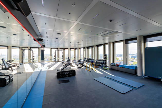 Berlin, 13.10.2026: Fitnessraum mit Hantelbänken, Kurzhanteln und Trainingsmatten im neuen DKB-Campus in der Europacity in Berlin-Mitte; durch die Fensterfront fällt der Blick über die Stadt. Die Deutsche Kreditbank AG (DKB) hat ihren neuen Hauptsitz heute offiziell eröffnet. Der von Kleihues + Kleihues entworfene, rund 82 Meter hohe Komplex besteht aus drei Gebäudeteilen mit fünf, elf und 19 Geschossen und rund 35.000 Quadratmetern Mietfläche; entwickelt wurde er von der CA Immo unter dem Projektnamen Upbeat. Rund 2.600 Mitarbeitende des DKB-Konzerns, die bislang auf mehrere Berliner Standorte verteilt waren, arbeiten nun gemeinsam im Campus. Die DKB ist Exklusivmieterin und hat einen Mietvertrag über 15 Jahre abgeschlossen.