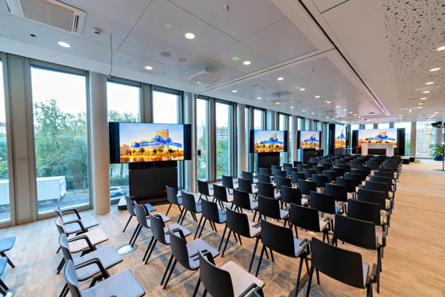 Berlin, 13.10.2026: Ein bestuhlter Veranstaltungssaal mit Bildschirmen, die das Motiv der Eröffnungswoche zeigen, im neuen DKB-Campus in der Europacity in Berlin-Mitte. Die Deutsche Kreditbank AG (DKB) hat ihren neuen Hauptsitz heute offiziell eröffnet. Der von Kleihues + Kleihues entworfene, rund 82 Meter hohe Komplex besteht aus drei Gebäudeteilen mit fünf, elf und 19 Geschossen und rund 35.000 Quadratmetern Mietfläche; entwickelt wurde er von der CA Immo unter dem Projektnamen Upbeat. Rund 2.600 Mitarbeitende des DKB-Konzerns, die bislang auf mehrere Berliner Standorte verteilt waren, arbeiten nun gemeinsam im Campus. Die DKB ist Exklusivmieterin und hat einen Mietvertrag über 15 Jahre abgeschlossen. Gastronomie-, Kultur- und