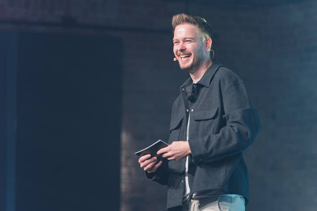 Berlin, October 16, 2026 Sascha Müller, host of NeulandPro Connect 2026, stands on stage in front of a dark brick wall, laughing with his cue ca