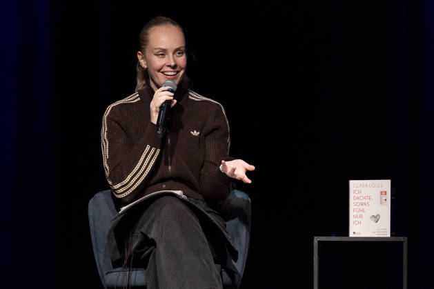 Poetry artist and author Clara LOESEL LÖSEL speaks to the audience with a smile during her reading—a preview show for her new book, *Ich dachte, sowas fühle nur ich*—seated with a microphone, in a single frame, a single photo, at the Theater am Tanzbrunnen, Cologne, on October 23, 2026