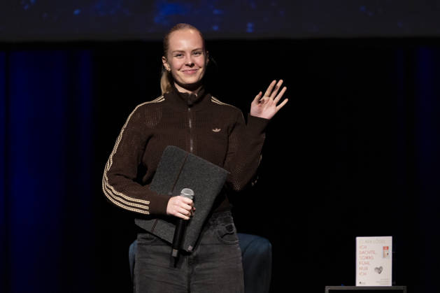 Poetry artist and author Clara LOESEL LÖSEL waves and smiles at the audience during her reading—a preview show for her new book, *Ich dachte, sowas fühle nur ich*—standing, single frame, single photo, at the Theater am Tanzbrunnen, Cologne, on October 23, 2026