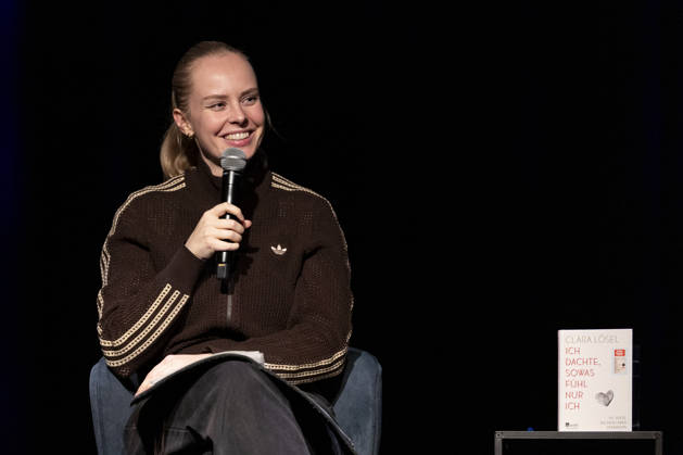 Poetry artist and author Clara LOESEL LÖSEL smiling, laughing, during her reading—a preview event for her new book, *Ich dachte, sowas fühle nur ich*—seated, holding a microphone; single frame, single photo, at the Theater am Tanzbrunnen, Cologne, on October 23, 2026