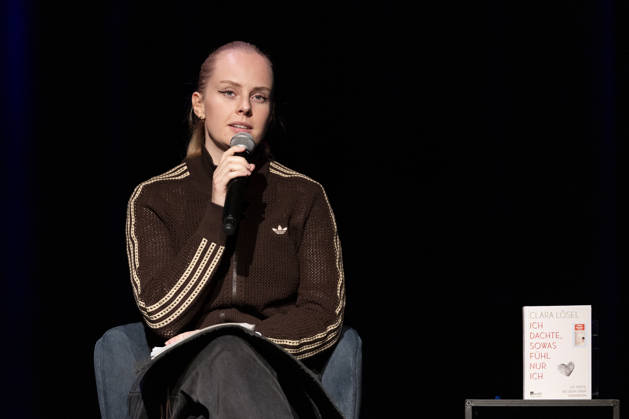 Poetry artist and author Clara LOESEL LÖSEL, serious and focused during her reading—a preview event for her new book, *Ich dachte, sowas fühle nur ich*—seated with a microphone; single frame, single photo, at the Theater am Tanzbrunnen, Cologne, on October 23, 2026