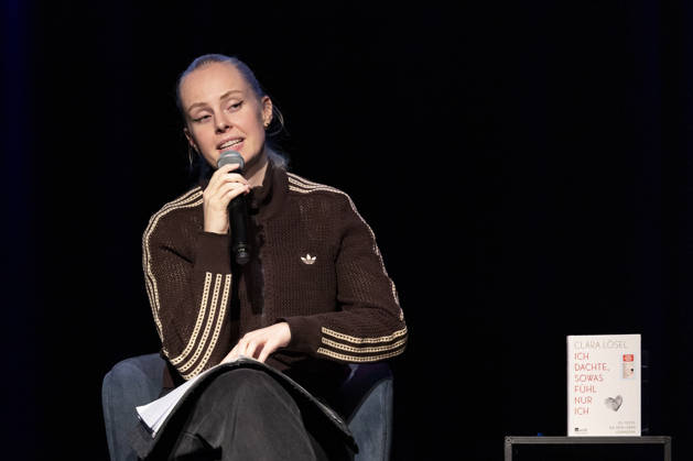 Poetry artist and author Clara LOESEL LÖSEL reading during her reading event, a preview show for her new book, *Ich dachte, sowas fühle nur ich* (I Thought I Was the Only One Who Felt That Way), seated with a microphone; single frame, single photo, at the Theater am Tanzbrunnen in Cologne on October 23, 2026