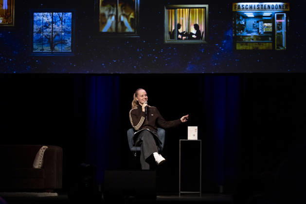 Poetry artist and author Clara LOESEL LÖSEL on stage during her reading, a preview show for her new book, *Ich dachte, sowas fühle nur ich* (I Thought I Was the Only One Who Felt That Way), seated with a microphone; single frame, single photo, at the Theater am Tanzbrunnen, Cologne, on October 23, 2026